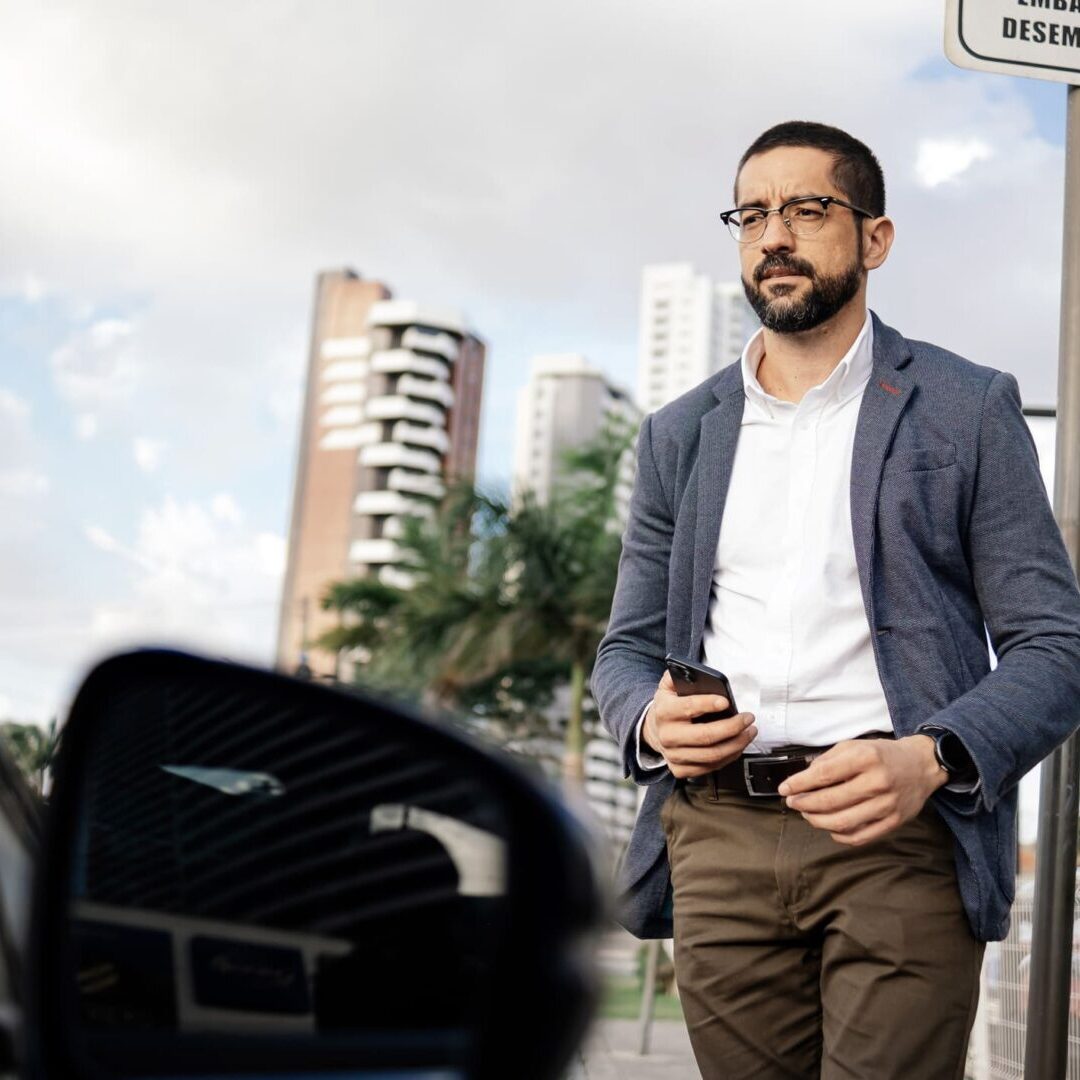 Adult business man using a smartphone while standing on an urban street with a car in the foreground. Concepts of ride hailing, transportation app, mobility and modern lifestyle