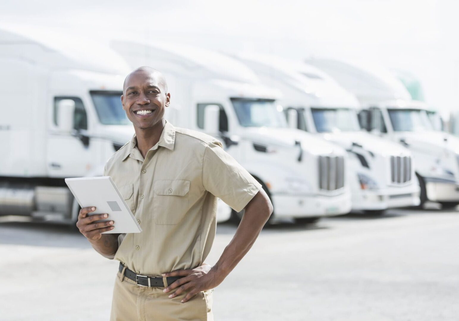 An African-American man in his 30s standing in front of a row of parked semi-trucks outside a distribution warehouse, holding a digital tablet.